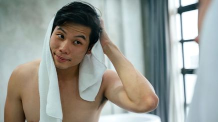 An Asian man rubbing a towel on his hair and looking at himself in the mirror