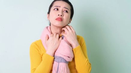 Asian woman wearing a yellow long-sleeved shirt and pink scarf scratching her neck.