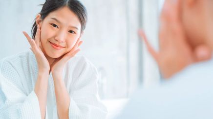 Smiling woman in white bathrobe touching her face while looking in the mirror.