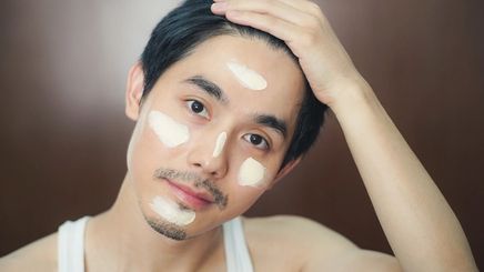 An Asian man with a mustache, beard, and face cream with his hand on his hair