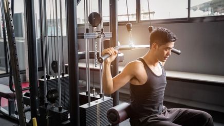 Young Asian man in a black sleeveless shirt holding a weights machine with both hands at the gym.