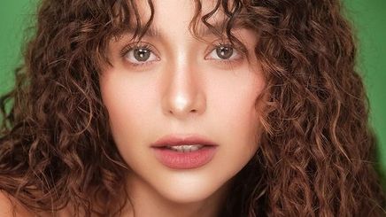 Yassi Pressman with curly hair and curtain bangs posing in front of the camera.