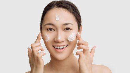 Smiling Asian woman applying moisturizer all over her face 