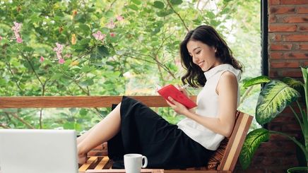 Asian woman sitting with a notebook on a balcony against outdoor greenery.
