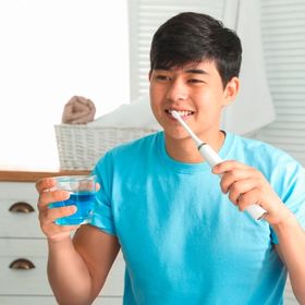 Asian man in blue brushing with an electric toothbrush and holding a cup of mouthwash.