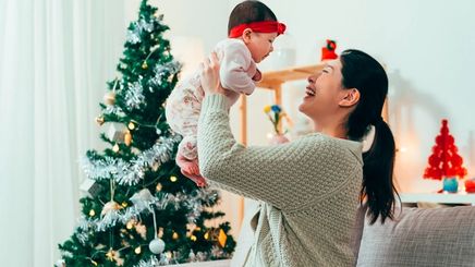 Asian mom carrying baby in front of Christmas tree.