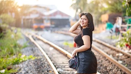 Full-figured Asian woman smiling and posing along train tracks.