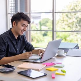 Young Filipino man sitting at desk with laptop, writing notes.