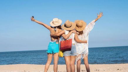 Three Asian women in hats and beach outfits facing the ocean at the beach.