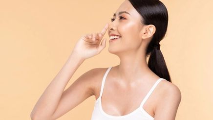 Side view of Asian woman smiling and touching her nose against a beige background.