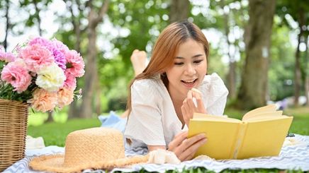 Asian woman having a solo picnic outdoors, reading a book and holding a macaron. 