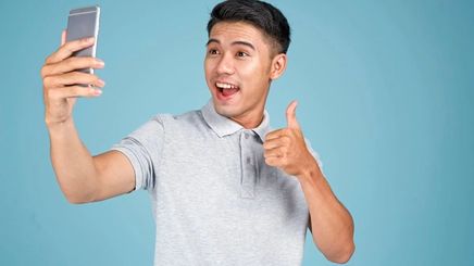 Young Asian man taking a photo with his phone giving the thumbs up against a blue background.