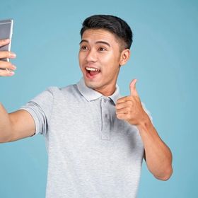 Young Asian man taking a photo with his phone giving the thumbs up against a blue background.