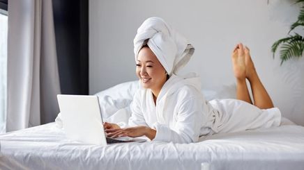 Asian man on bed with towel around head and smiling at laptop