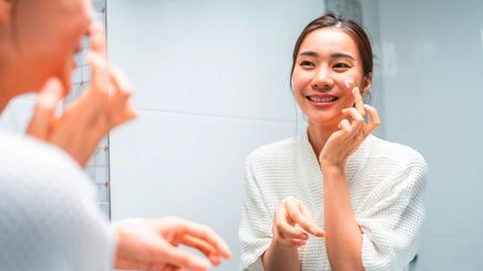 Asian woman applying moisturizer on her cheek in front of a mirror.