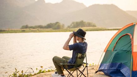 Back view of a relaxed man with his head resting on his arms, looking at a sunset and a view of a lake with a backpack and tent.