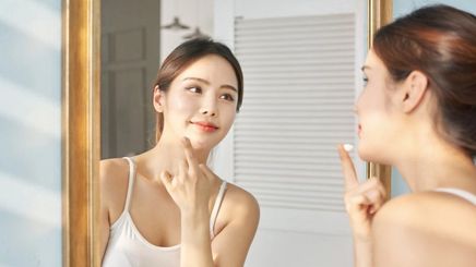 Asian woman applying product while looking in a mirror. 