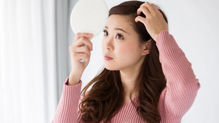 Woman wearing pink blouse checking her scalp in a handheld mirror. 