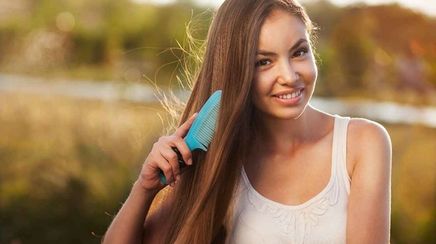 Woman in white tank top brushing her long hair with blue comb.