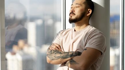 Man with tattoos on his arm closes his eyes while leaning against a pillar