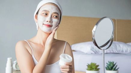Asian woman applying a cream mask on her face in front of a mirror.