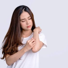 Young Asian woman in white scratching her forearm against a white background.