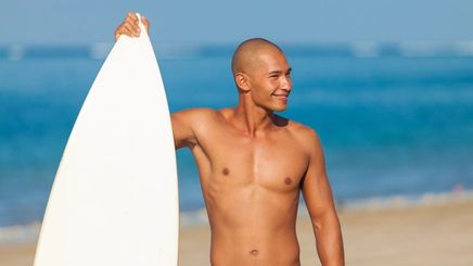 Young man with a shaved head holding a surfboard on the beach