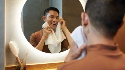 Asian man holding a face towel, staring at his reflection in the mirror. 