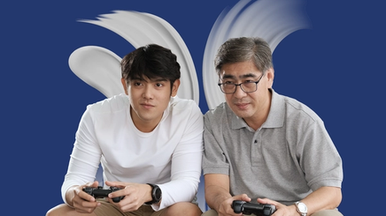 An Asian father and son sitting on a couch, playing video games 