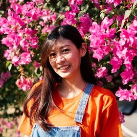 Young Asian woman in an orange shirt posing with flowers in the background.