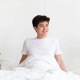 Smiling Asian man in bed with white bedsheets and visible plants.