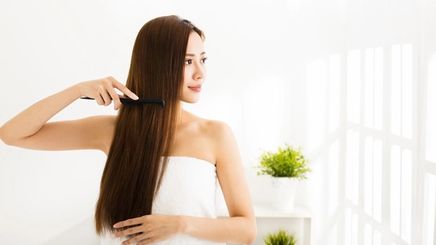 Side view of Asian woman smiling combing her long, straight hair in a white, bright room.
