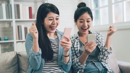 Two Asian women sitting on the couch, laughing while looking at their cellphones
