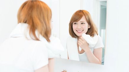 An Asian woman with short hair smiling at a mirror and touching her face 