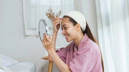 Asian teen applying toner with a cotton pad while holding a mirror. 