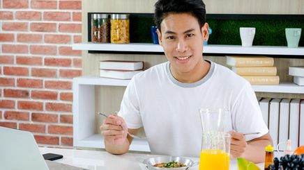Young Asian man eating a healthy breakfast while looking at his laptop