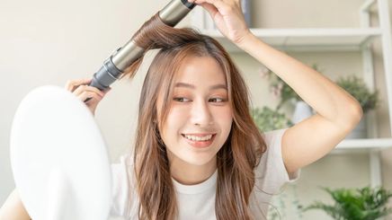Asian woman curling her hair with hot iron in front of a mirror.
