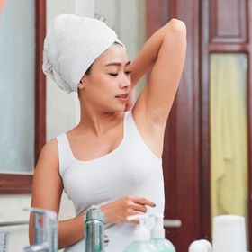Asian woman in white and wearing towel while looking at underarm.