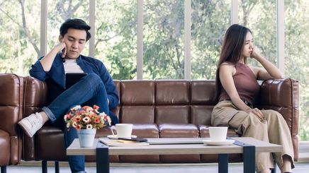 Asian man and woman sitting on opposite ends of a brown leather couch against a background of a glass window with trees visible.