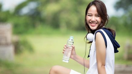 Asian woman smiling holding a bottle of water and exercising outdoors. 