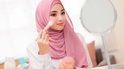 Muslim woman applying blush on her cheek with a fluffy brush.