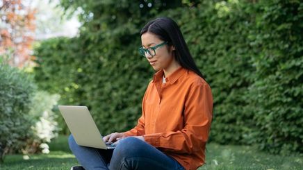 An Asian woman sitting on the grass, working on her laptop