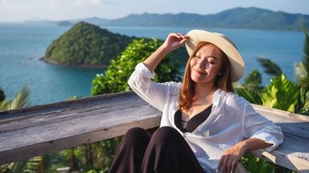 Asian woman wearing a hat, sitting on a deck overlooking the ocean. 