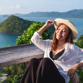 Asian woman wearing a hat, sitting on a deck overlooking the ocean. 