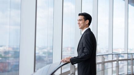 Asian man in a suit putting his hand on a railing and looking out the window of a building.