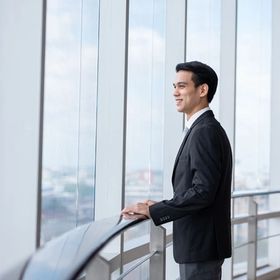 Asian man in a suit putting his hand on a railing and looking out the window of a building.