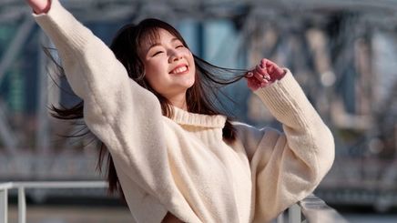 Smiling woman in cropped white sweater with long black hair raising her arms in the air.