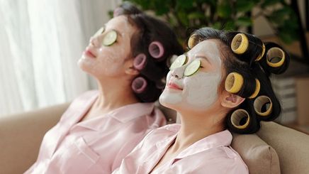 Two Asian women having a spa day with mud masks, cucumber on their eyes, and rollers in their hair.