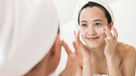 Asian woman in a bathroom applying face cream in front of a mirror. 