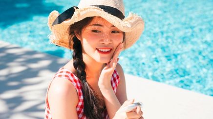 Woman in sun hat applying sunscreen onto her face by the pool.